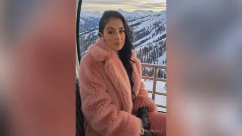 Young woman in a cozy pink faux fur coat enjoying a scenic snowy mountain view from a ski lift gondola.