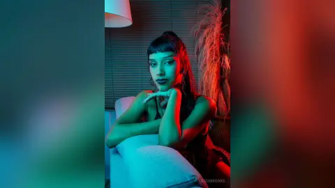 LilithBrooks rests her chin on her hands while posing in a dark room with blue and red lighting.