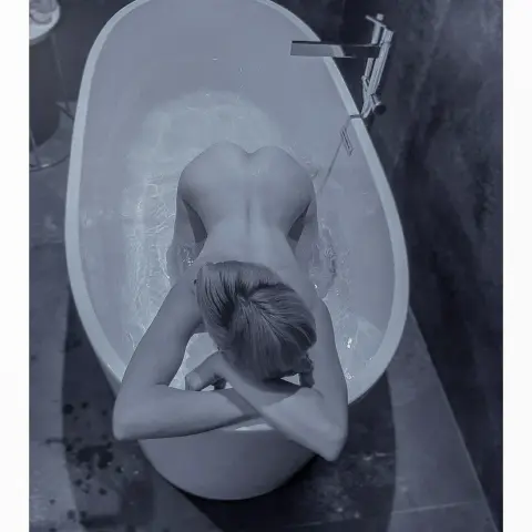 Nude woman relaxing in a white freestanding bathtub in a moody, modern dark bathroom setting.