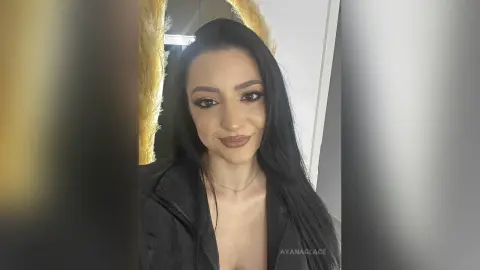 Smiling woman with long black hair and smoky eye makeup wearing a black jacket in a bright indoor setting.