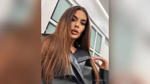 Glamorous woman with long brown hair wearing a black leather jacket in a bright modern interior.