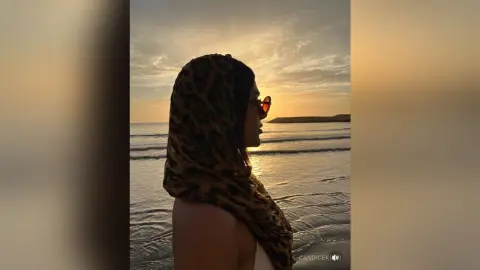 Woman in leopard print headscarf and sunglasses gazing at the ocean during a peaceful golden hour sunset.