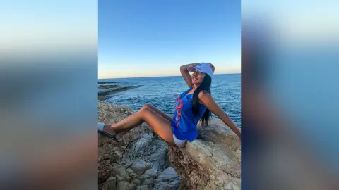 MissyJolie sits on a rock overlooking the ocean wearing a blue tank top and white bottoms with a white sun visor.
