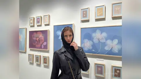Woman in a black leather jacket and headscarf standing in a modern art gallery surrounded by floral paintings.