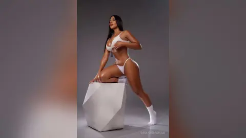 CassandraDuque poses in a white bikini and white socks, leaning against a white geometric block against a grey background.