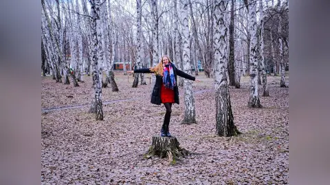 Blonde woman in a black coat and red dress standing on a tree stump in a birch forest during autumn.