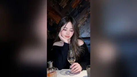 Young woman in a black off-the-shoulder top enjoying wine at a dimly lit restaurant.