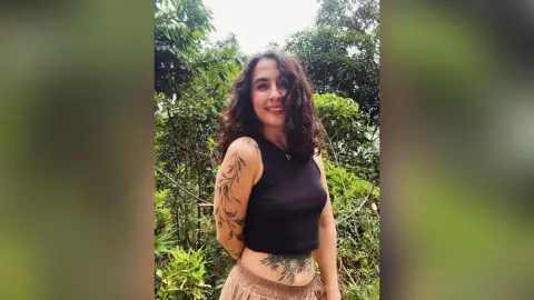 RossiNoleen smiles in a black tank top and tan skirt, standing outdoors against a background of green trees and leaves.