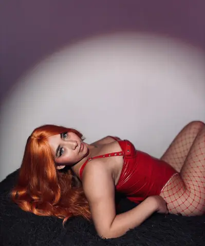 Redheaded woman in a red bodysuit and fishnet tights lying on a black rug in moody studio lighting.