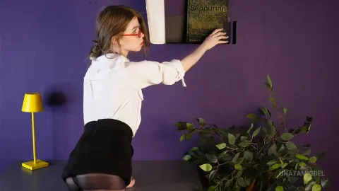 Young woman in a white blouse and black skirt reaching for a book on a shelf against a purple wall.