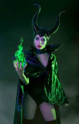 Woman in Maleficent cosplay with black horns and flowing cape posing in mystical green magical smoke.