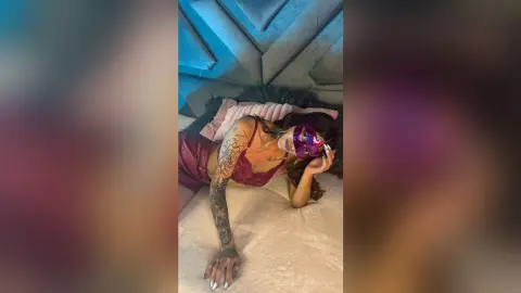 Tattooed woman in a burgundy lace outfit and purple masquerade mask lounging on a bed in moody lighting.