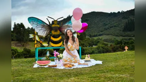 Cheerful woman in a white floral dress celebrating a birthday picnic with cake and balloons in a green meadow.