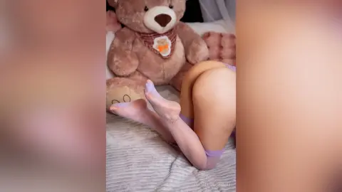 Woman in white sheer tights relaxing on a bed next to a large brown teddy bear in a cozy bedroom setting.