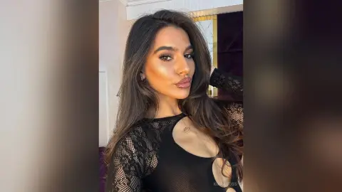 Glamorous woman with long brown hair wearing a black lace cutout bodysuit in a soft indoor setting.