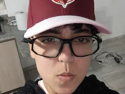 Young man with glasses and a maroon baseball cap posing in a casual indoor setting.