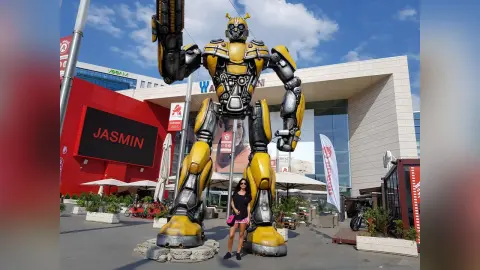 Woman in black outfit standing next to a giant Transformers Bumblebee statue at a sunny outdoor shopping mall.