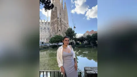 Smiling woman in a grey dress and white shirt standing by a lake with Sagrada Familia in Barcelona.