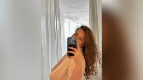 Young woman in a white blouse taking a selfie in a bright, airy room with sheer white curtains.