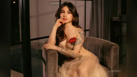 Elegant woman in a sheer beige tulle gown sitting on a velvet armchair in a moody, dimly lit room.