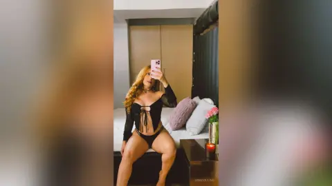 Blonde woman in a black long-sleeve crop top and bikini bottoms posing for a mirror selfie in a cozy bedroom.