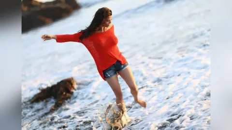 Cheerful woman in a red sweater and denim shorts playing in ocean waves on a sunny beach.