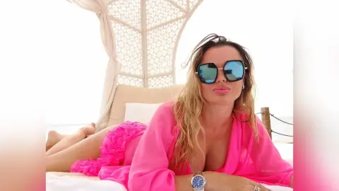 MilenaBia reclines on a white bed under a canopy, wearing a bright pink robe and blue-tinted sunglasses.