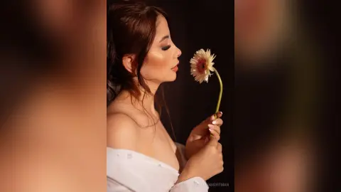ValentinaHoffman poses in profile, holding a single flower near her face while wearing a white off-the-shoulder top.