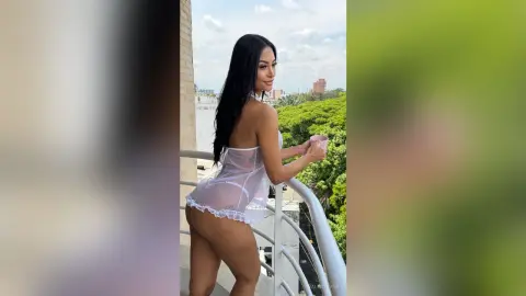 Young woman in a white lace negligee holding a pink mug on a sunny balcony overlooking a city skyline.