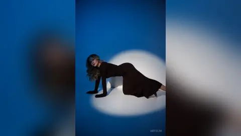 Elegant woman in a long brown dress posing against a blue studio background with dramatic spotlighting.