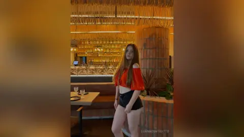 LuanaContratto poses in a red off-the-shoulder crop top and black shorts, standing in a warm, lit restaurant setting with long brown hair.