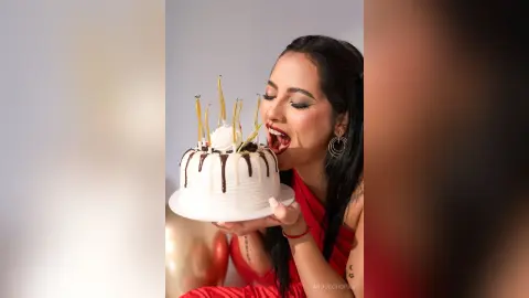 Cheerful woman in a red dress celebrating a birthday with a white frosted cake and lit candles.