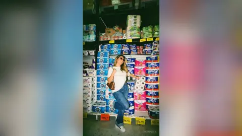 Smiling woman in white t-shirt and blue jeans posing in a grocery store aisle filled with paper towel rolls.