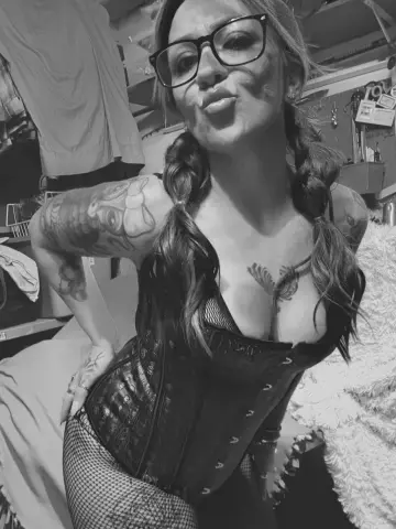 NovaBRollin pouts her lips while wearing a black corset and fishnet top, showing off her tattoos and pigtails in this black and white photo.