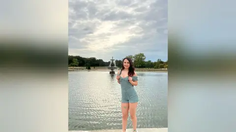 RossiNoleen stands in front of a large fountain wearing a light blue off-the-shoulder romper.