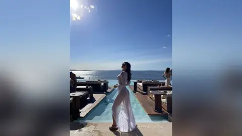 Woman in a flowing white dress standing by a luxury rooftop pool under a bright sunny sky.