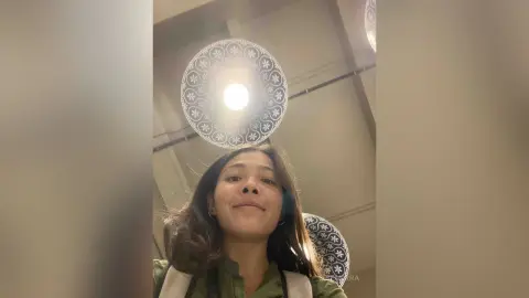 Smiling young woman in a green shirt relaxing under decorative patterned ceiling lights in a cozy indoor setting.