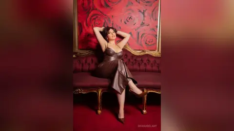 Elegant woman in a brown satin dress sitting on a vintage velvet sofa against a red floral painting background.