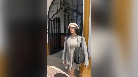 Young woman in a grey plaid blazer and beige beret standing by an ornate black gate in soft daylight.