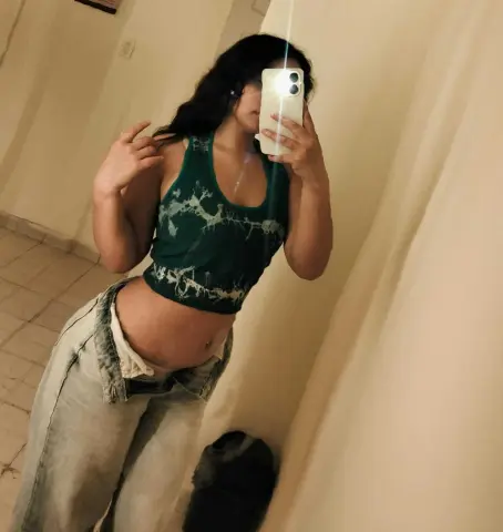 Young woman in a green tie-dye crop top and baggy jeans taking a casual mirror selfie in a bright room.