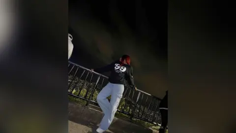 Young woman in a black graphic sweatshirt and light blue jeans posing on a balcony at night.