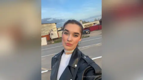 EmelyMastorovo poses outdoors wearing a black leather jacket over a white top with a silver choker necklace.