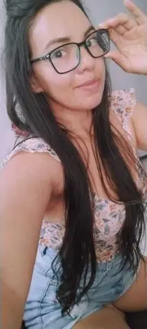 Smiling woman with long dark hair wearing glasses and a floral print top in a bright indoor setting.