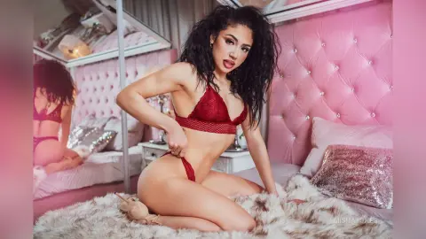 Glamorous woman in red lace lingerie posing on a pink tufted bed in a dreamy, feminine bedroom setting.