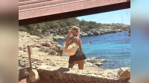 Young woman in a black summer dress holding a straw bag by the turquoise ocean on a sunny rocky coastline.