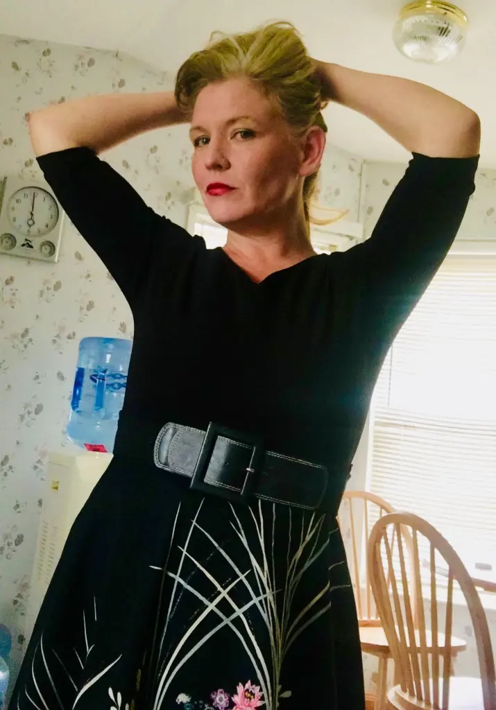 CoraElaine, a blonde woman, looks at the camera while wearing a black t-shirt against a light-colored curtain background.