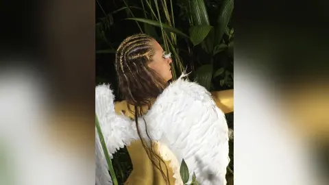 AndreaKalo poses outdoors in a jungle setting, wearing white angel wings and a yellow top with her hair in long blonde braids.