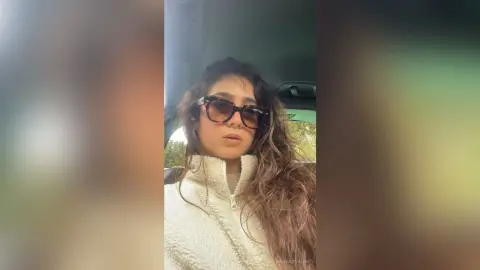 Young woman with long wavy hair wearing sunglasses and a white fleece jacket sitting in a car.