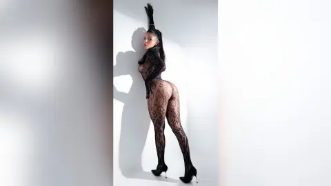 Elegant woman in a black lace bodysuit and sheer tights posing against a white wall in a moody studio setting.