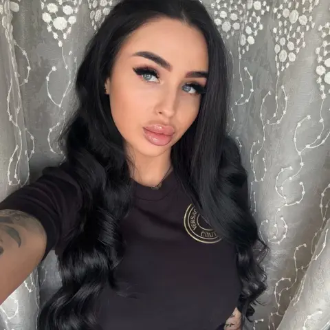 Glamorous woman with long wavy hair wearing a black Versace t-shirt posing against a lace curtain backdrop.